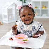 A happy baby sits in a highchair using the Bumkins Silicone First Feeding Set: Minnie Mouse Light Pink while wearing a matching bib.