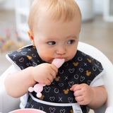 A baby with a patterned bib curiously chews a spoon from Bumkins Silicone First Feeding Set: Minnie Mouse Light Pink, looking aside.