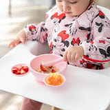 A baby in a bib self-feeds oatmeal and strawberries from Bumkins Silicone First Feeding Set: Minnie Mouse Light Pink at a white highchair.