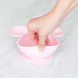 A hand holds a Bumkins Silicone First Feeding Set: Minnie Mouse Light Pink, demonstrating the suction cup bottom on a marble surface.