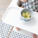 A baby in a checkered bib sits in a high chair, eating from a Bumkins Silicone First Feeding Set: Gray with a checkered mat on the floor.