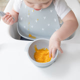 A girl in a gray bib in a high chair eats orange puree with a bowl and spoon from Bumkins Baby-Led Weaning Essential 6-Piece Set: Gray.