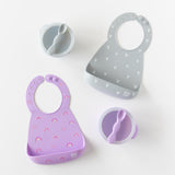 Bumkins Silicone Bib 2 Pack: Hearts and Rainbows lays flat on a white background next to gray and purple bowl & spoon sets.