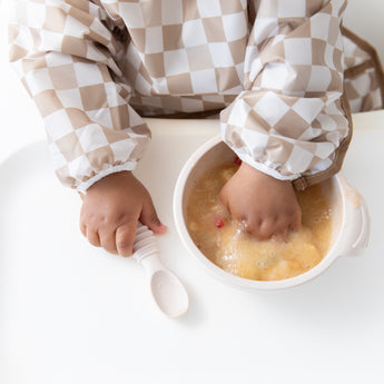 Close-up of a baby in a checkered bib, eating applesauce with their hand from Bumkins Silicone First Feeding Set: Sand on a high chair tray.