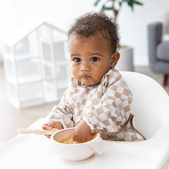 A baby in a high chair, wearing a Bumkins Sleeved Bib 2 Pack: Palm Check, eats from a beige bowl in a bright room.