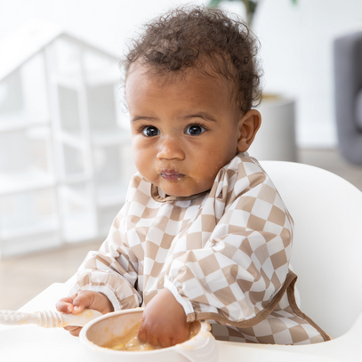 A baby in a high chair, wearing a checkered bib, uses Bumkins Silicone First Feeding Set: Sand in a bright room.