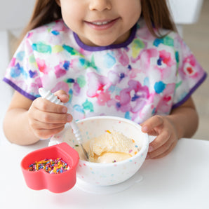 A child in a floral bib beams while enjoying ice cream from a Bumkins Silicone First Feeding Set: Vanilla Sprinkle  at a white table.