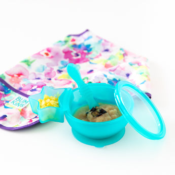 Bumkins Silicone First Feeding Set: Blue Jelly with applesauce, sits next to a floral bib and a blue star-shaped cup on a white background.