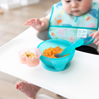 A baby sits in a high chair with a Bumkins Silicone First Feeding Set: Blue Jelly filled with pureed sweet potatoes near a pink cup of fruit.