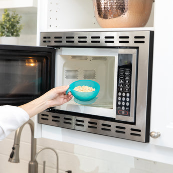 A person places a Bumkins Silicone First Feeding Set: Blue Jelly with oatmeal into an open microwave.