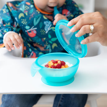 A child sits in a high chair chewing a spoon as an adult hand lifts the lid of a Bumkins Silicone First Feeding Set: Blue Jelly with fruit.