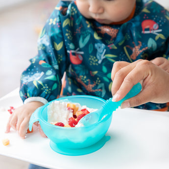A toddler in a blue bib enjoys yogurt and fruit from a Bumkins Silicone First Feeding Set: Blue Jelly at a high chair with an adult's help.