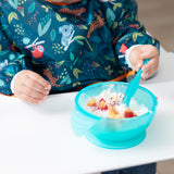 Close-up of a child in a colorful bib eating fruit and yogurt from a Bumkins Silicone First Feeding Set: Blue Jelly on a white high chair tray.