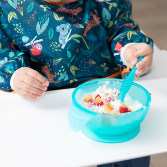 Close-up of a child in a colorful bib eating fruit and yogurt from a Bumkins Silicone First Feeding Set: Blue Jelly on a white high chair tray.