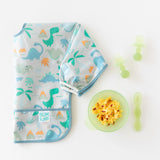 Bumkins Silicone First Feeding Set: Green Jelly with eggs sits next to a folded dinosaur bib & green toddler utensils on a white background.