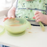 Close-up of a baby in a high chair wearing a green bib, eating puree from a Bumkins Silicone First Feeding Set: Green Jelly.