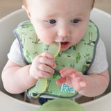 A baby in a high chair wearing a green bib chews the spoon of a Bumkins Silicone First Feeding Set: Green Jelly.