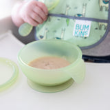 A baby holds a spoon with a Bumkins Silicone First Feeding Set: Green Jelly on a high chair tray while a green bib peeks behind.