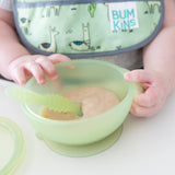 Close-up of a baby holding a Bumkins Silicone First Feeding Set: Green Jelly with applesauce, wearing a bib with animal designs.
