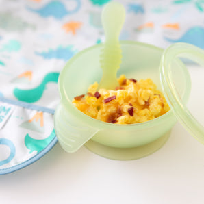 Close-up of Bumkins Silicone First Feeding Set: Green Jelly filled with scrambled eggs & bacon next to a dinosaur bib on a white background.