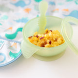 Close-up of Bumkins Silicone First Feeding Set: Green Jelly filled with scrambled eggs & bacon next to a dinosaur bib on a white background.