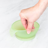 A hand holds a Bumkins Silicone First Feeding Set: Green Jelly, demonstrating the suction cup bottom on a marble surface.