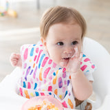 In a high chair, a baby with a colorful bib enjoys messy mealtime using Bumkins Silicone First Feeding Set: Pink Jelly.