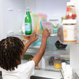 A child places a Bumkins Silicone Flat Reusable Bag 2 Pack, Pink into an open refrigerator next to other bags and a bottle of sparkling water.