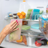 A hand places a Bumkins Silicone Flat Reusable Bag 2 Pack, Pink among lettuce, fruits, and containers into the open fridge.