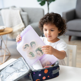 A curly-haired child packs food into a space-themed lunchbox with Bumkins Silicone Flat Reusable Bag 2 Pack, Lavender in a bright room.