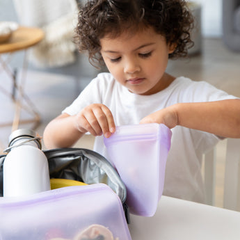 A child places food in a Bumkins Silicone Flat Reusable Bag 2 Pack, Lavender, with a lunchbox and water bottle on the table nearby