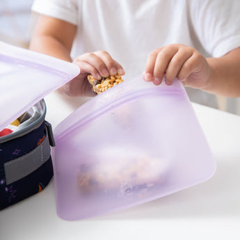 A child's hands place a granola bar in a Bumkins Silicone Flat Reusable Bag 2 Pack, Lavender near a lunchbox on a white table.