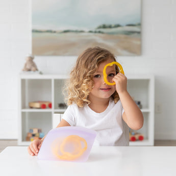 A child peeks through a yellow bell pepper slice at a white table with Bumkins Silicone Flat Reusable Bag 2 Pack, Lavender.