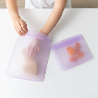 A child places a sandwich in a Bumkins Silicone Flat Reusable Bag 2 Pack, Lavender, with fruit in the other beside it, on a white table.