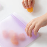 Close-up of a child's hands placing an apricot into a Bumkins Silicone Flat Reusable Bag 2 Pack, Lavender on a white table.