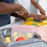 Two people chop veggies in a kitchen while a Bumkins Silicone Flat Reusable Bag 2 Pack, Pink with cucumber slices sits in an open lunchbox.