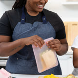 A person in blue apron places a sandwich into a Bumkins Silicone Flat Reusable Bag 2 Pack, Pink in a bright kitchen.