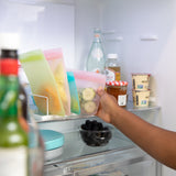 A person stores a Bumkins Silicone Flat Reusable Bag 2 Pack, Pink with cucumbers in an open fridge filled with drinks and berries.