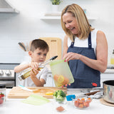 A woman holds a Bumkins Silicone Flat Reusable Bag 2 Pack, Sage with salmon as a boy pours oil, surrounded by fresh ingredients in a kitchen.
