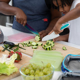 Two people chop cucumbers, surrounded by grapes, veggies, and Bumkins Silicone Flat Reusable Bag 2 Pack, Sage nearby on a kitchen counter.