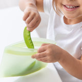 A child smiling in a white shirt, placing peas into a Bumkins Silicone Flat Reusable Bag 2 Pack, Sage at a white table.