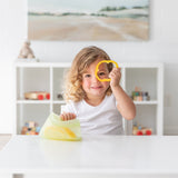 A child playfully holds a bell pepper slice over one eye at a white table with a Bumkins Silicone Flat Reusable Bag 2 Pack, Sage.