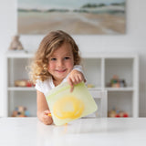 A child smiles at a white table holding a Bumkins Silicone Flat Reusable Bag 2 Pack, Sage, with shelves in the background.