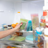 A hand places a Bumkins Silicone Flat Reusable Bag 2 Pack, Sage filled with veggies in a fridge, with other bags, a milk jug & fruit visible.