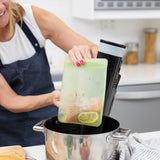 A person in an apron places a sealed Bumkins Silicone Flat Reusable Bag 2 Pack, Sage with salmon and lemon into a pot with a sous vide device.