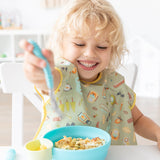 A curly blonde child in a green bib smiles, enjoying a meal with Bumkins Grip Bowl Essential 4-Piece Mealtime Set: Blue at a white table.