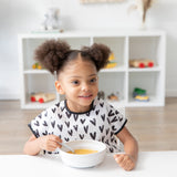 A girl with curly hair wears a black & white bib and eats soup with Bumkins Grip Bowl Essential 4-Piece Mealtime Set: Marble at a table.