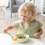 A blonde child in a green bib eats yogurt, berries, & granola with Bumkins Grip Bowl Essential 4-Piece Mealtime Set: Sage at a white table.