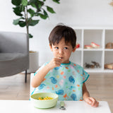 A young boy in a blue bib eats a meal with Bumkins Grip Bowl Essential 4-Piece Mealtime Set: Sage at a white table.