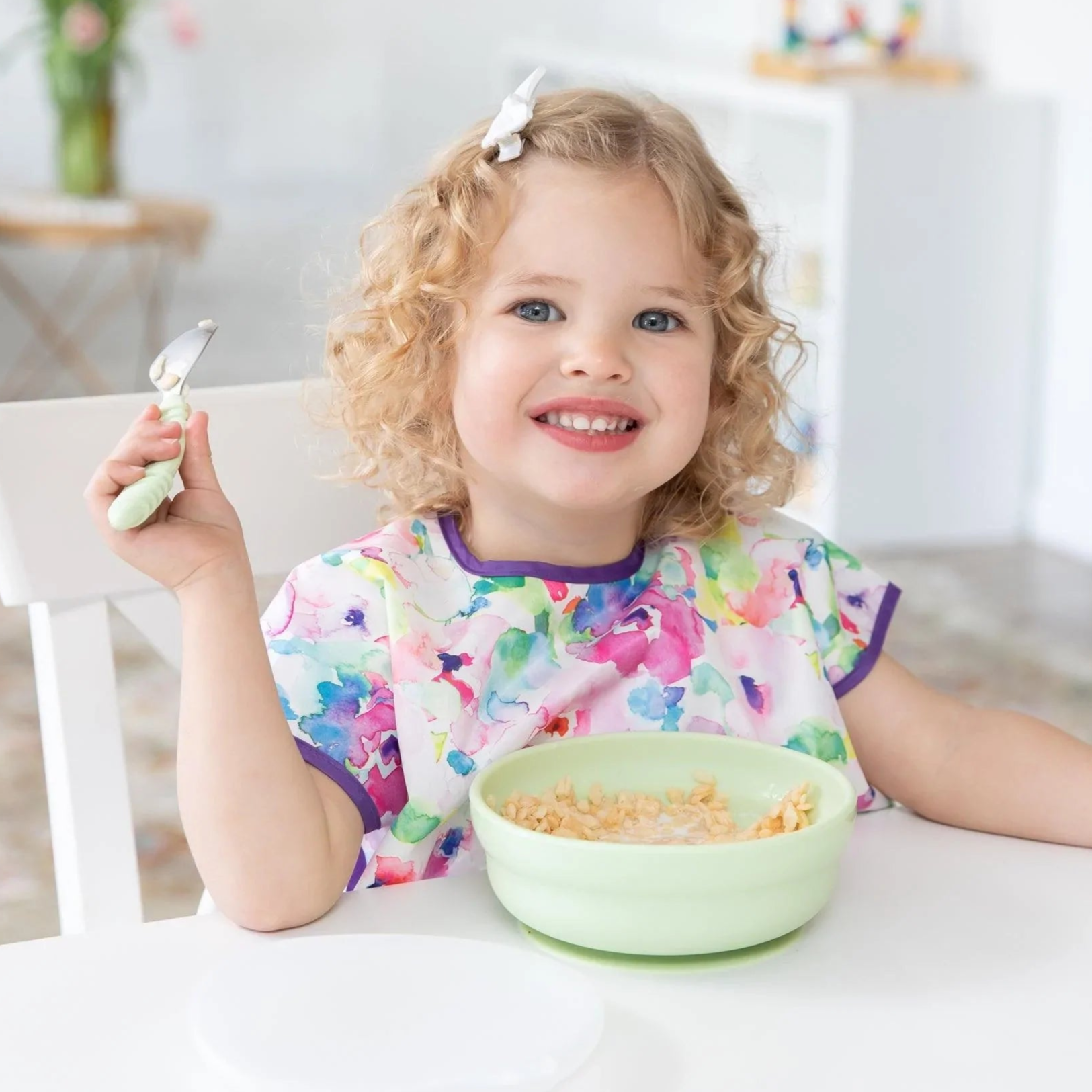 A curly-haired child beams, enjoying cereal from a Bumkins Silicone Grip Bowl: Sage, adding charm with their bright bib and bow.